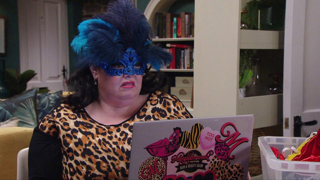 Mandy in a blue feathered mask sitting at a computer and gesticulating in Emmerdale